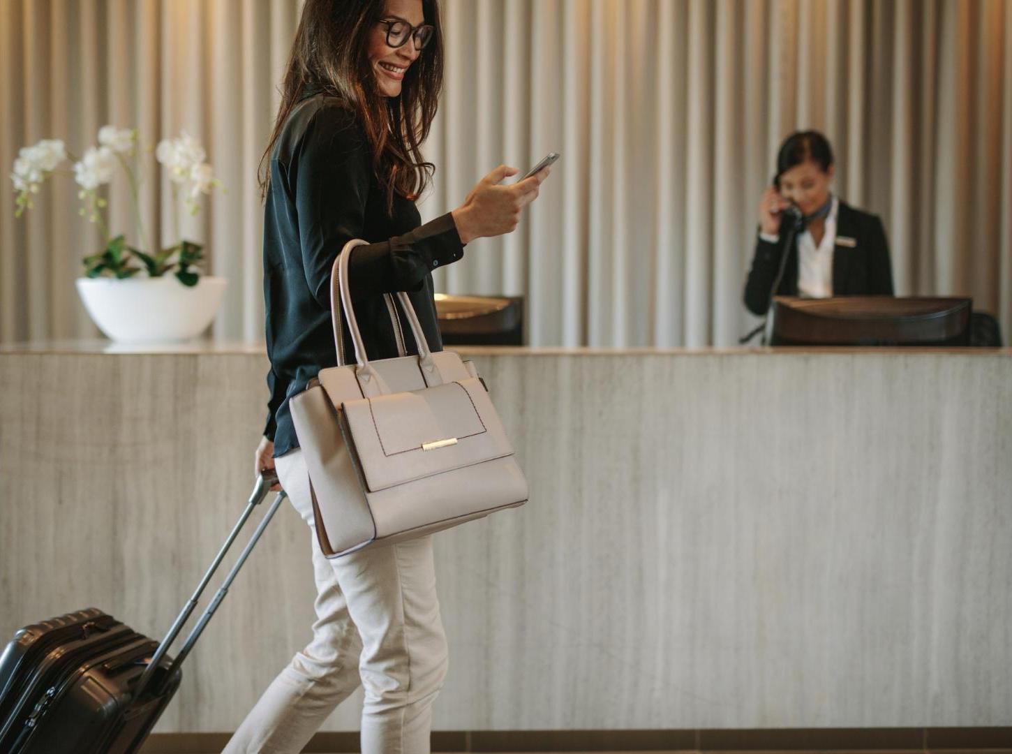 Business-woman-pull-her-luggage-and-smile-to-her-phone-in-front-of-hotel-front-desk-and-receptionist-behind-her-call-the-other-guest