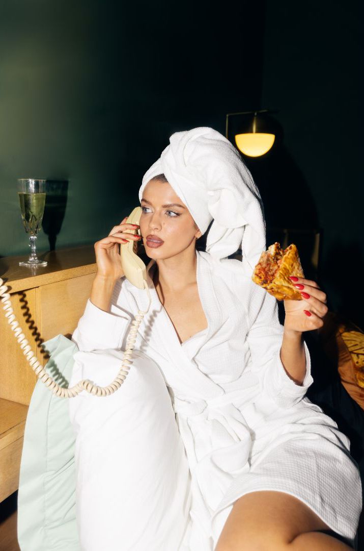 A woman wearing a white bathrob and red brown lip gloss and fresh clean manicure nails with red color is eating peperoni pizza with extra cheese while sitting on a white bed accompanied by champagne.