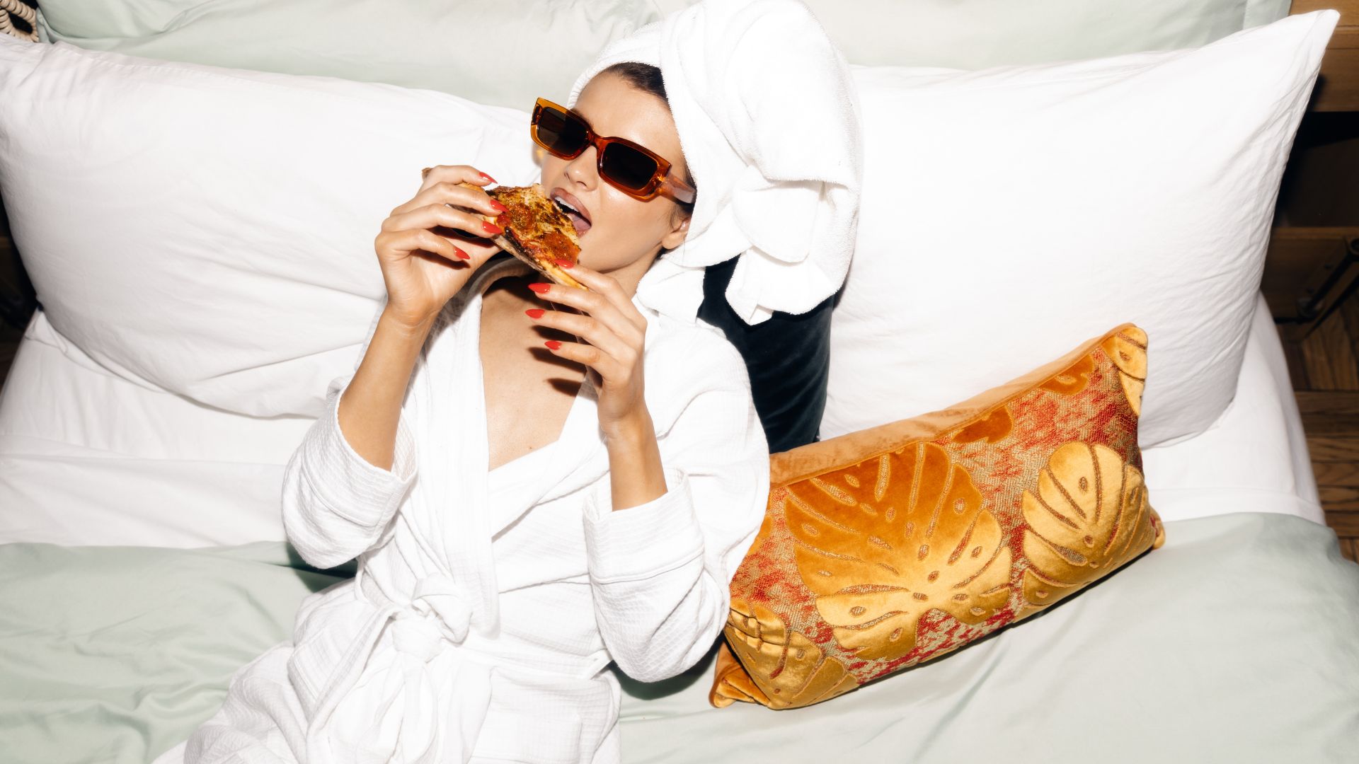 A woman wearing a bathrob and leopard brown aesthetic glasses for women is eating giant pieces of pizza while lying on her white and comfortable mattress next to her is a golden orange pillow with leaves on it