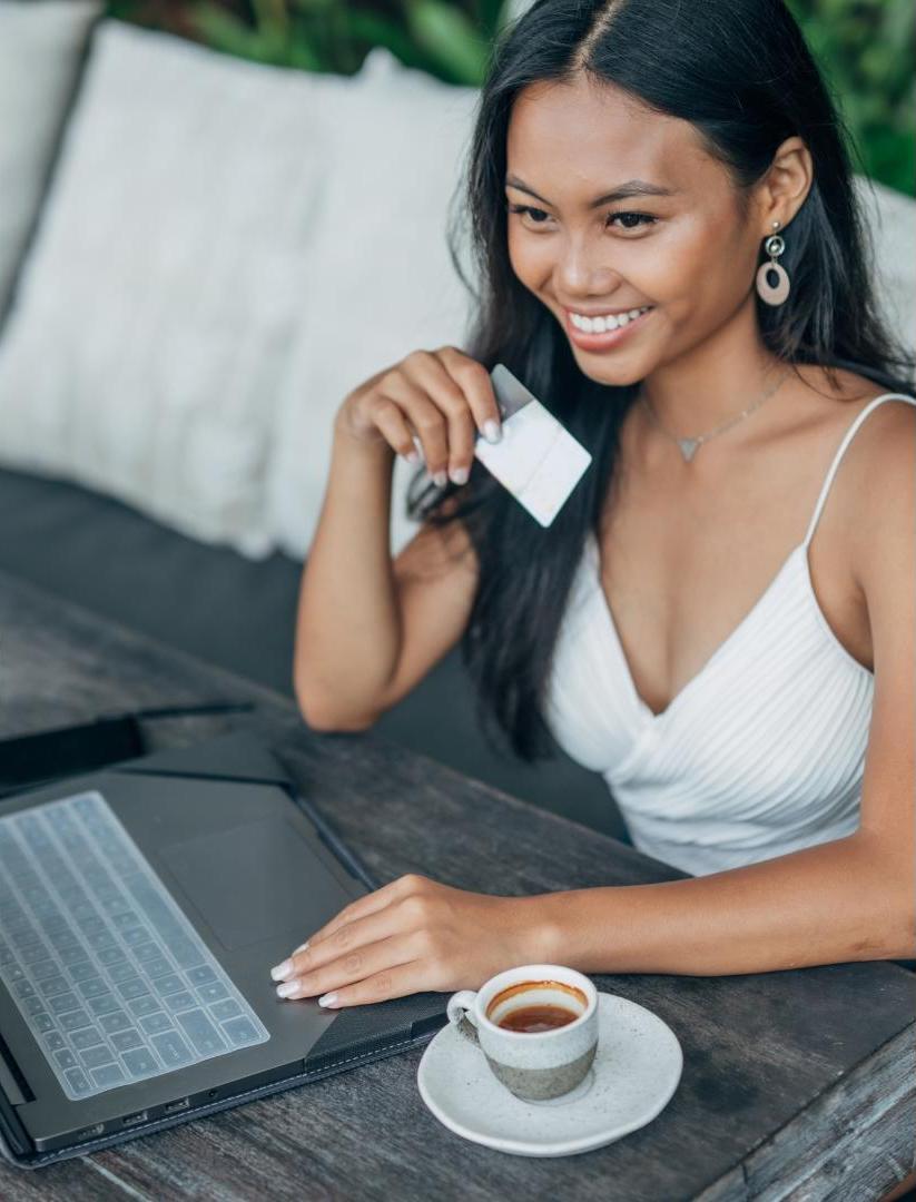 Indonesia-pretty-female-model-in-white-dress-smile-and-hold-the-debit-card-with-laptop-in-front-of-her-and-a-cup-of-coffee-symbolic-of-hotel-guest-persona
