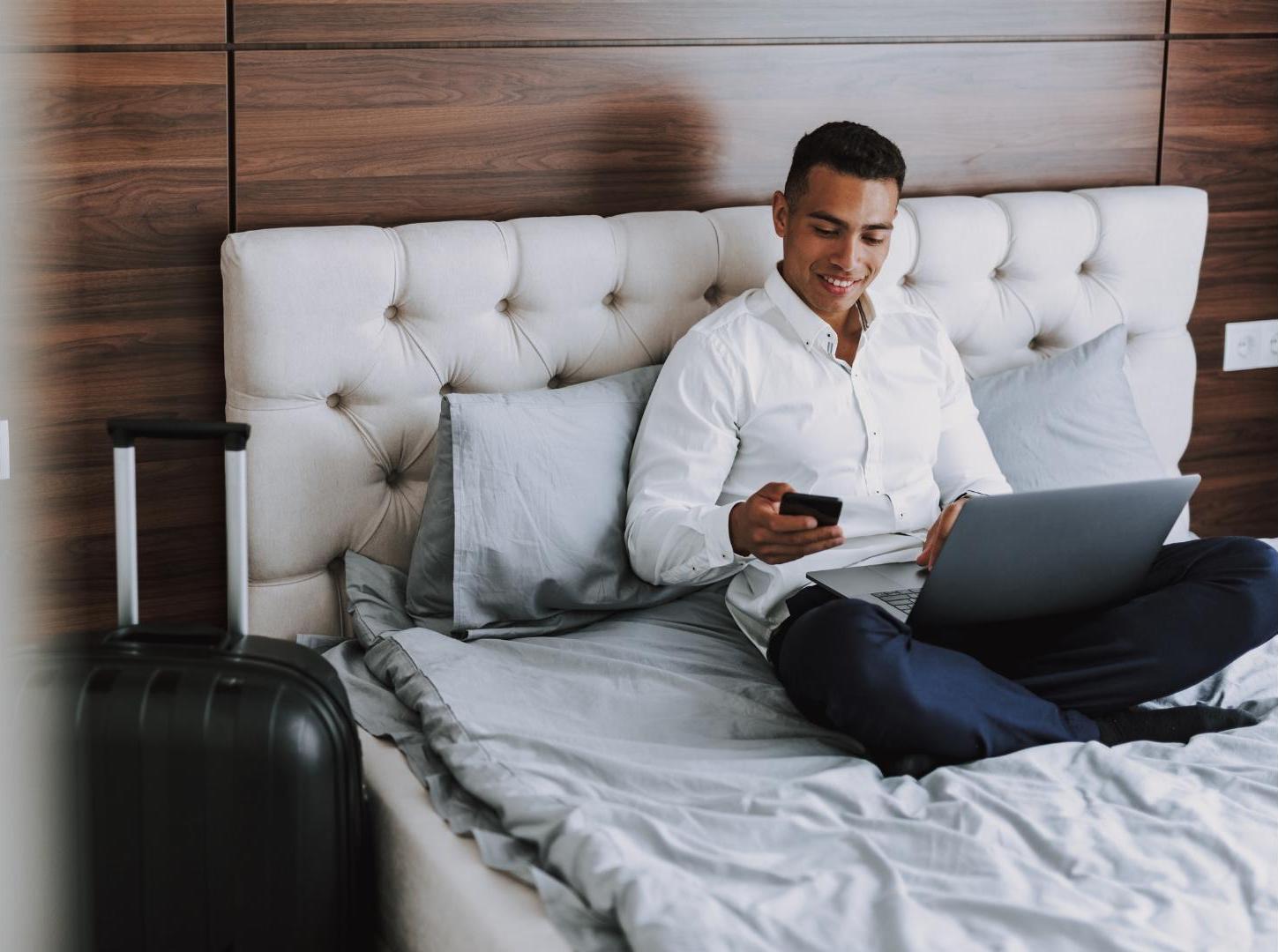 Latin-male-model-with-white-shirt-and-dark-blue-long-pants-smiling-to-his-phone-on-the-hotel-bad-with-macbook-on-his-two-legs-and-luggage-next-to-it