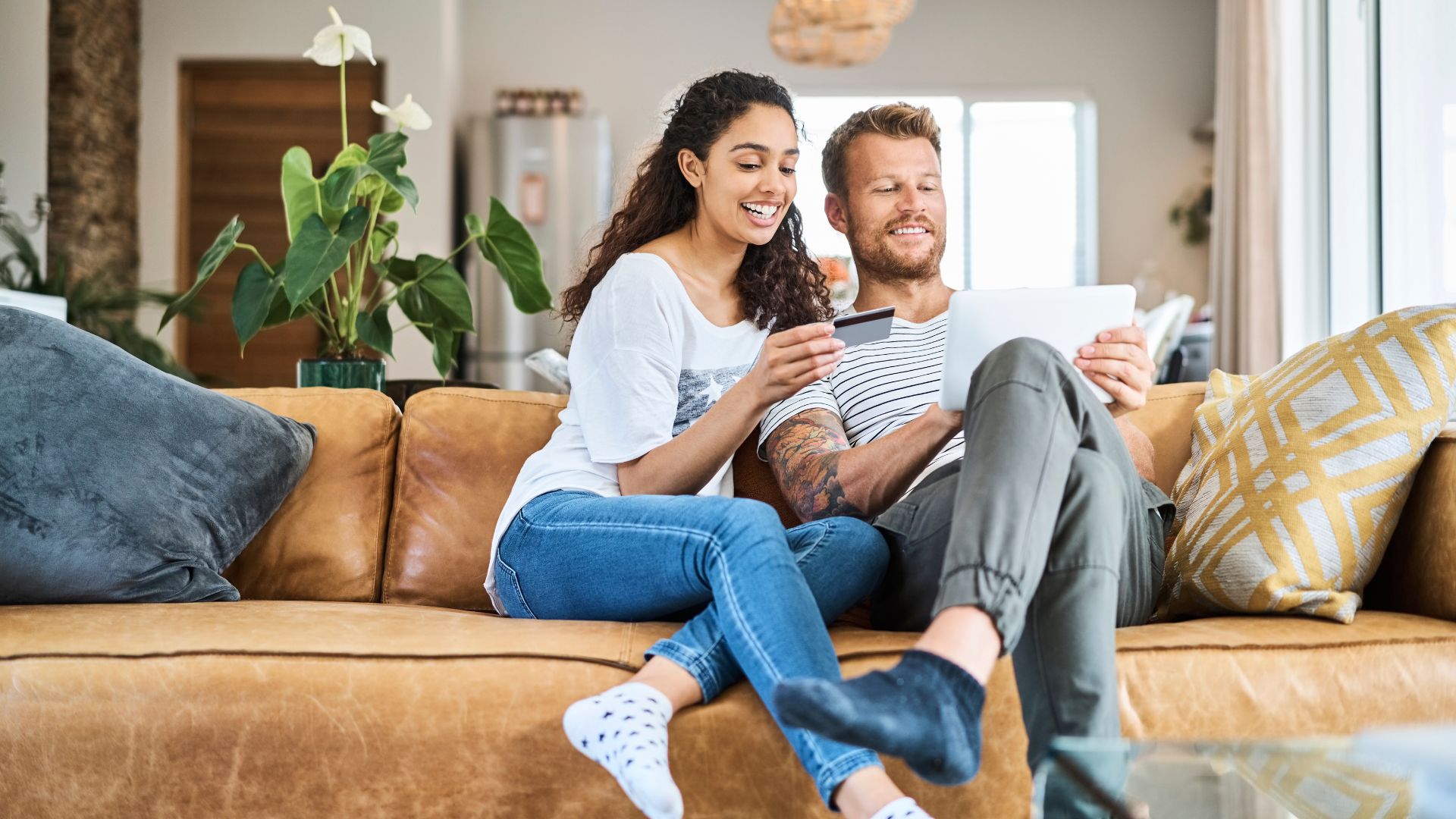 A woman wearing a white T-shirt and skinny jeans with curly hair is sitting on a brown leather sofa with aesthetic design pillows with a man wearing a white T-shirt with black stripes and gray jogger pants is looking to make a transaction on the marketplace using a credit card in the living room
