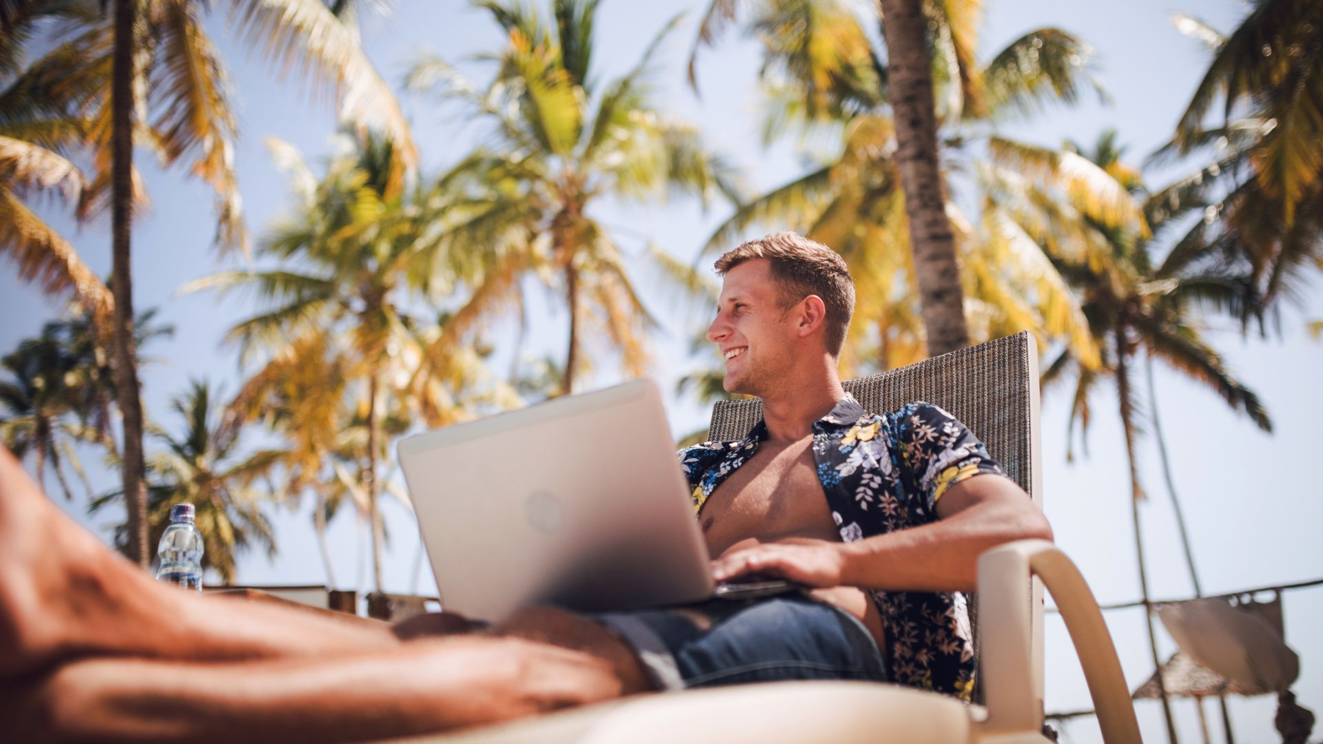 A man with a muscular body sitting on a sunbed wearing a floral Balinese shirt and knee-length shorts is working to create a memorable brand identity under the hot sun by the pool
