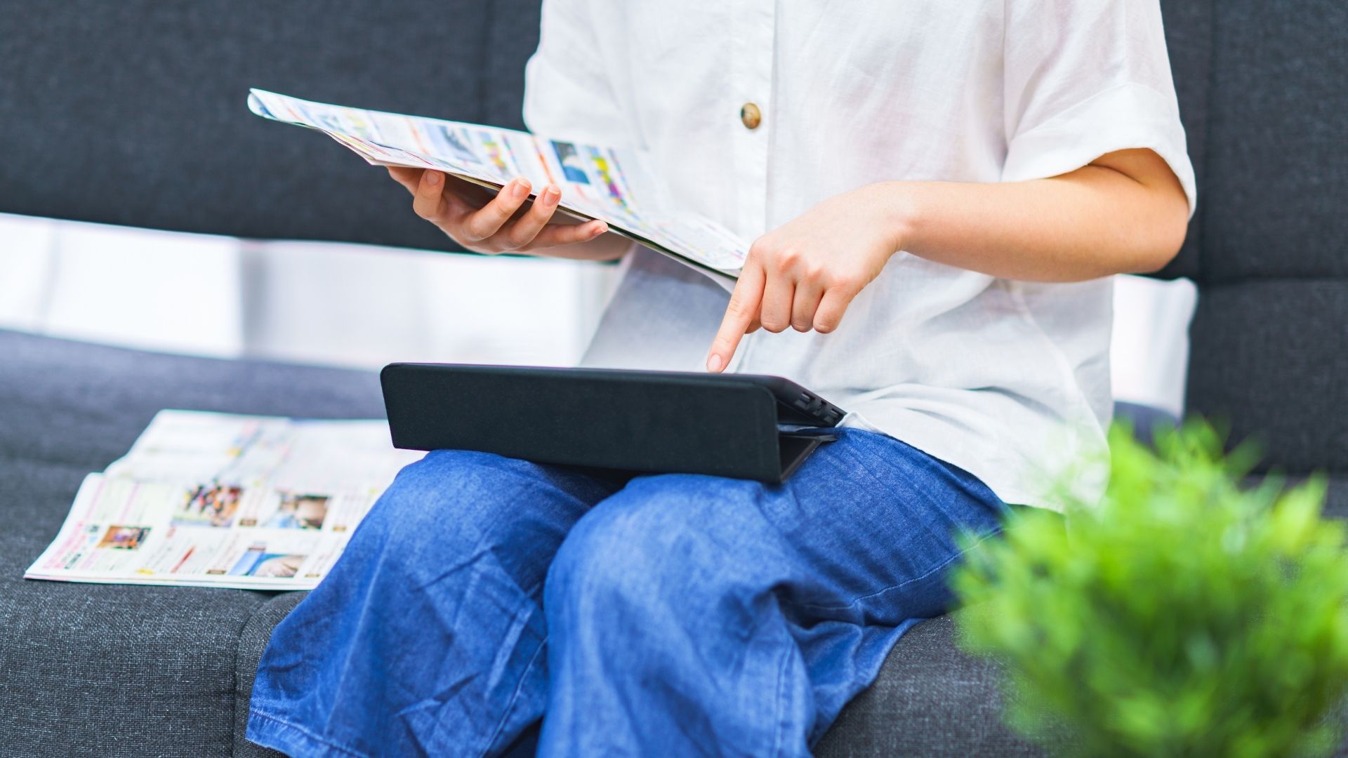 A woman wearing a white shirt with long jean pants is searching for relevant keywords through a black ipad and with a source from a fashion magazine.