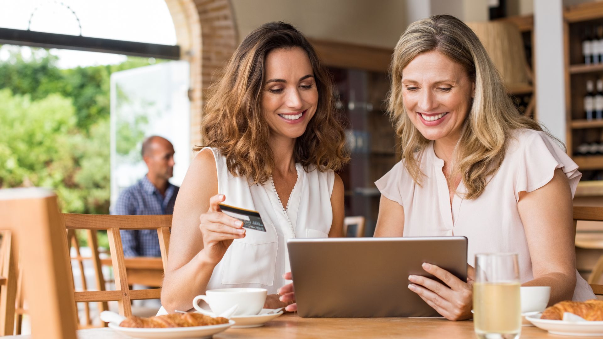 Two women wearing white blouses working together at an aesthetic café in Bali while having breakfast croissants and hot latte cafes want to make payments using credit cards via ipad
