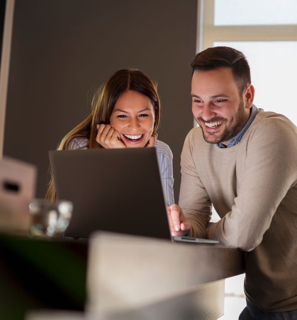 A woman wearing a white blue stripe shirt and a man wearing a blue shirt layered with a brown sweater are laughing and smiling happily because they see the amazing user experience of a website