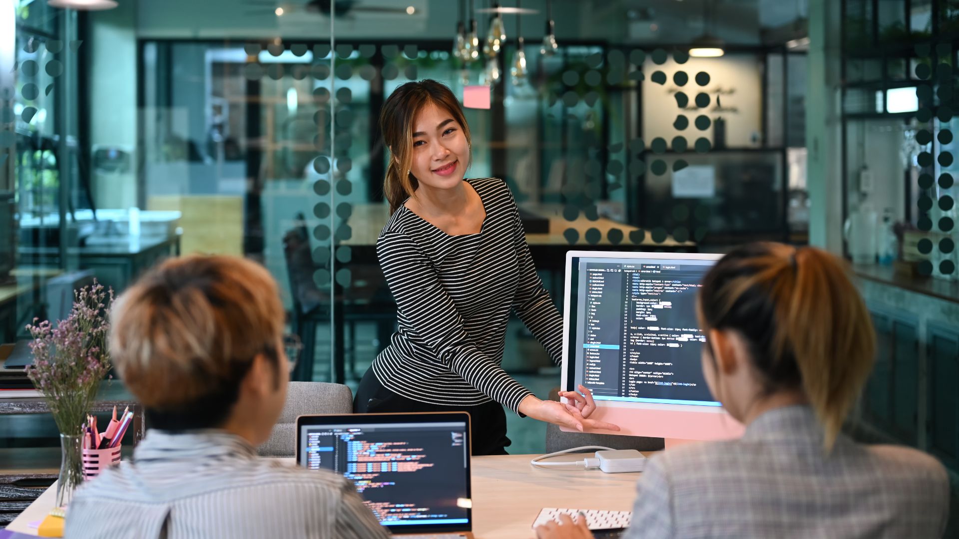 A woman wearing a black and white striped long-sleeved shirt is presenting about the web development team's performance using a Mac monitor in an office space located in a high-rise building.