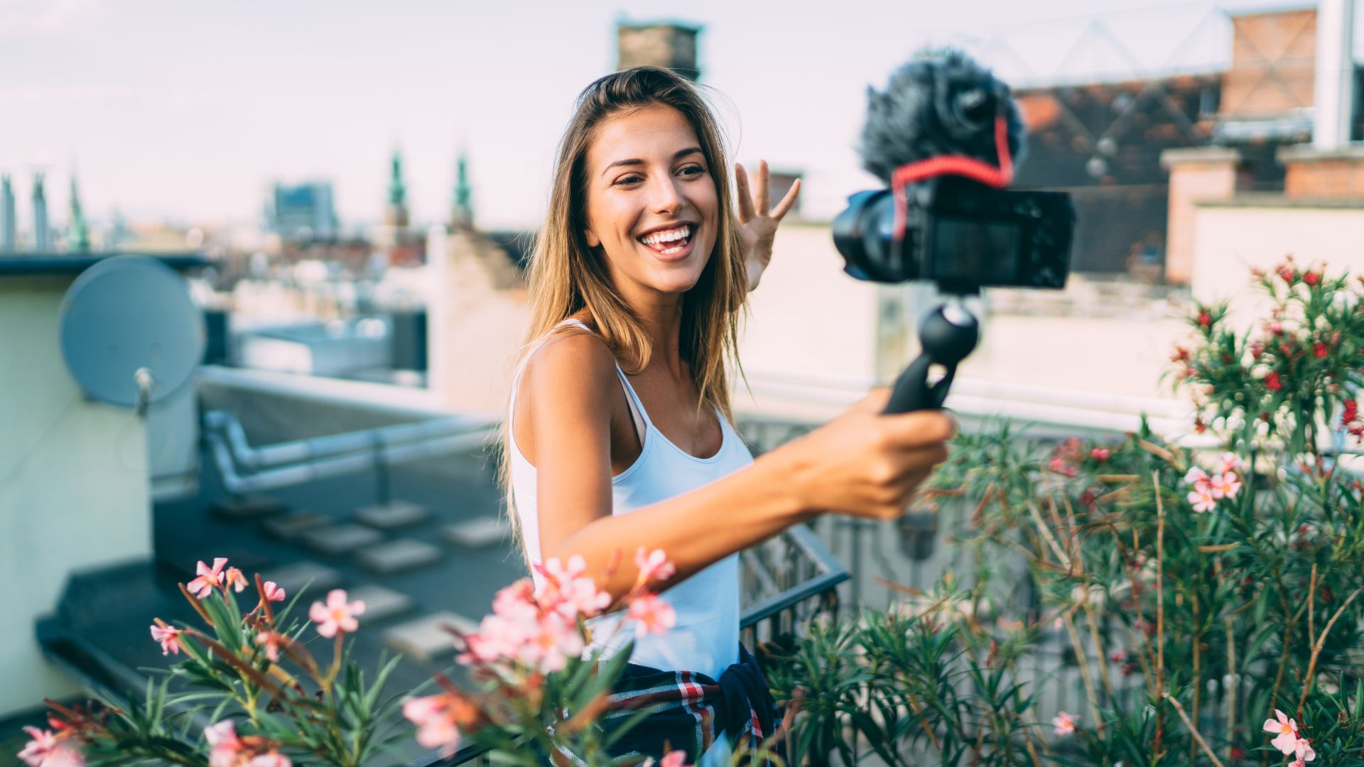 A female KOL influencer wearing a white tanktop is creating content with her camera that has a top quality camera mic on a rooftop with a flower garden.