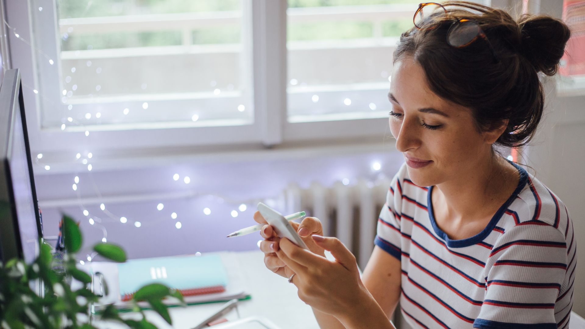 A woman wearing a red and blue striped white t-shirt with a messy bun hairstyle is doing social media management using her cell phone in her aesthetic room decorated with white LED lights.
