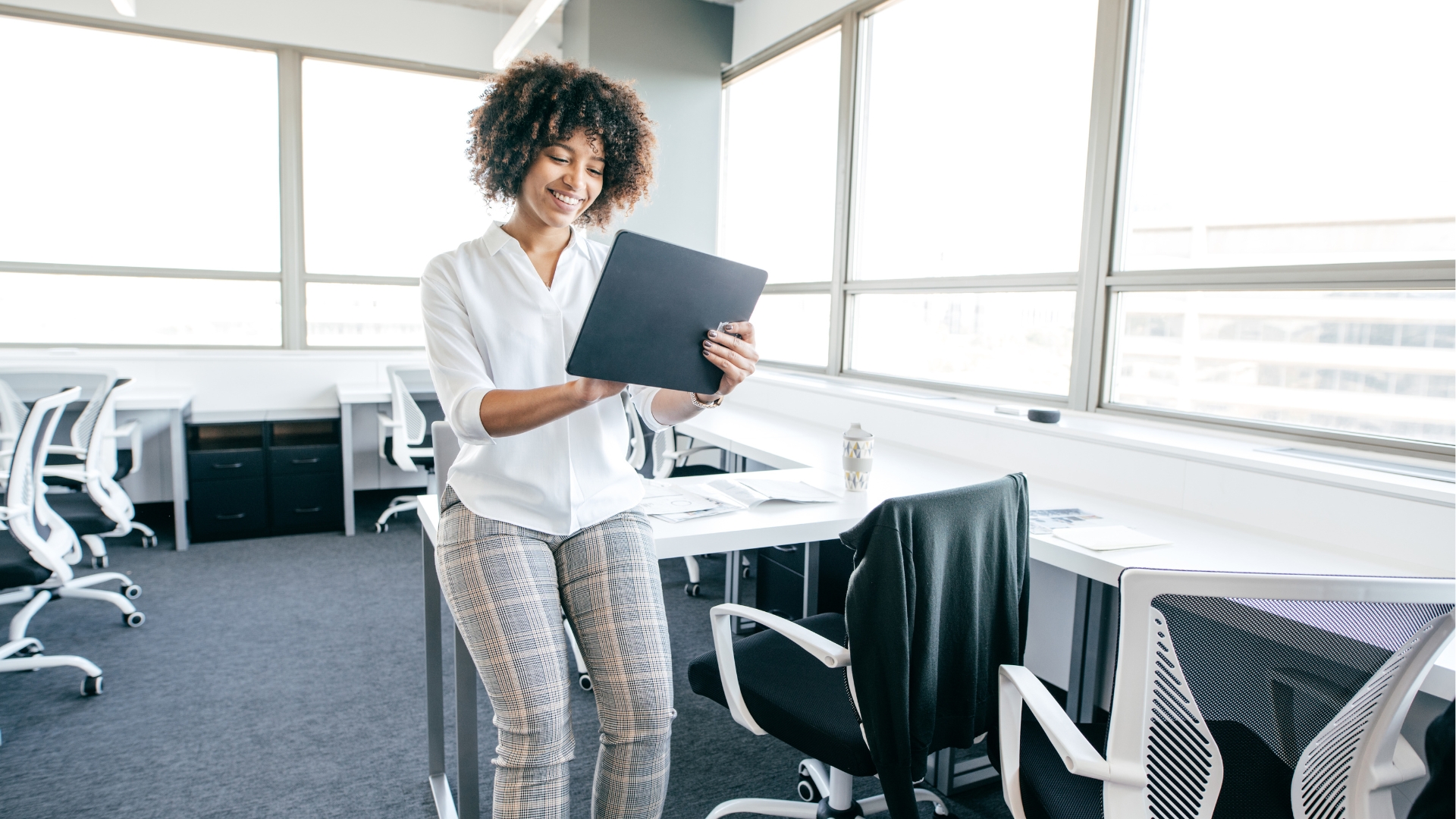 A woman with curly hair wearing a plain white shirt and patterned fabric pants is sitting at her office desk holding an ipad doing personalized account management.