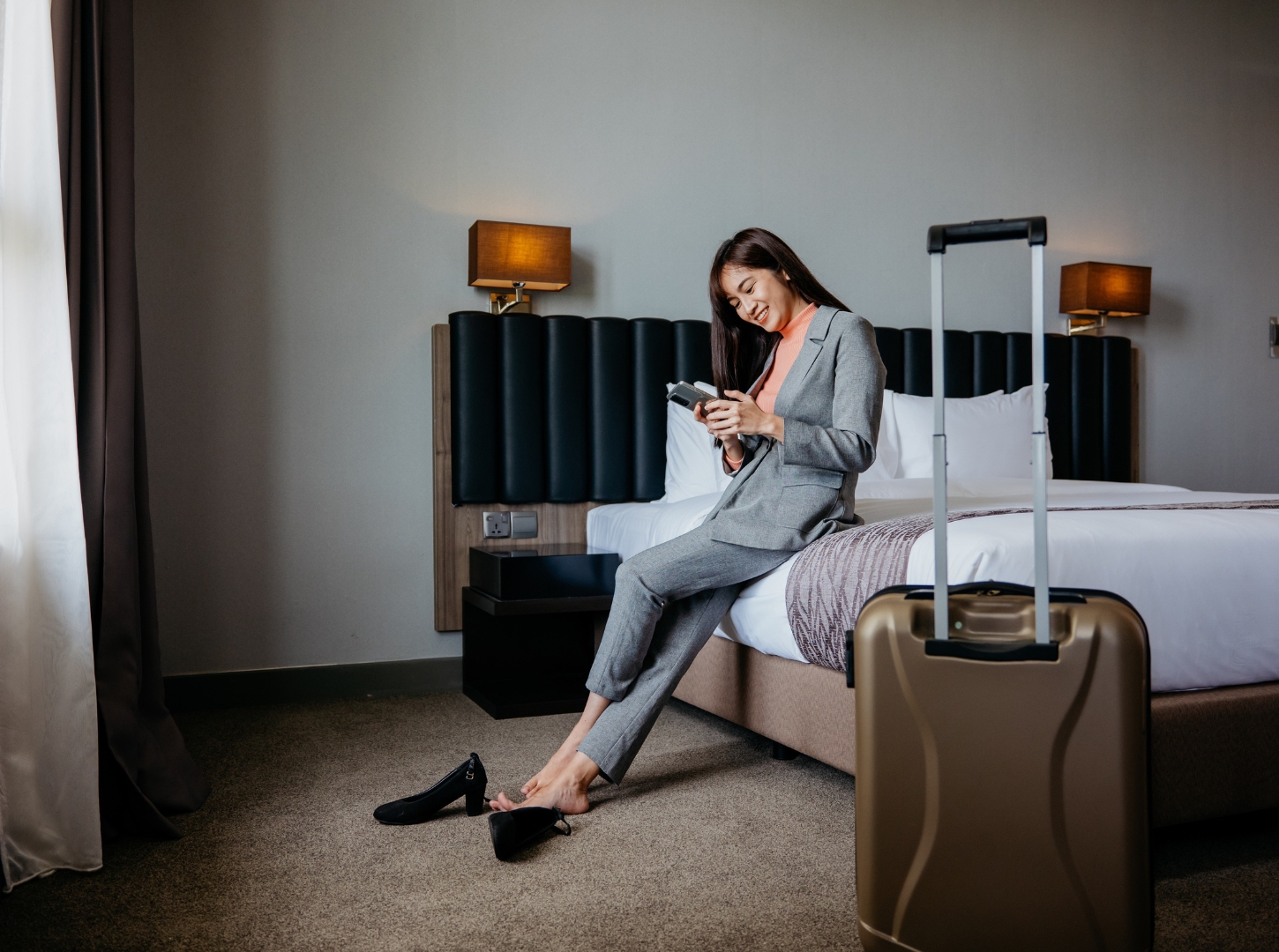 Woman hotel guest in grey sit in suit the hotel bedroom look at her smartphone with brow luggage in front of her