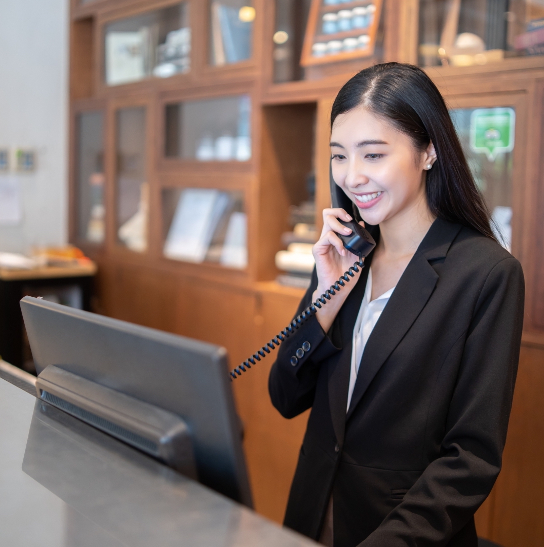a female hotel receptionist with black hair and black suit pick the phone up and the black computer in front of her