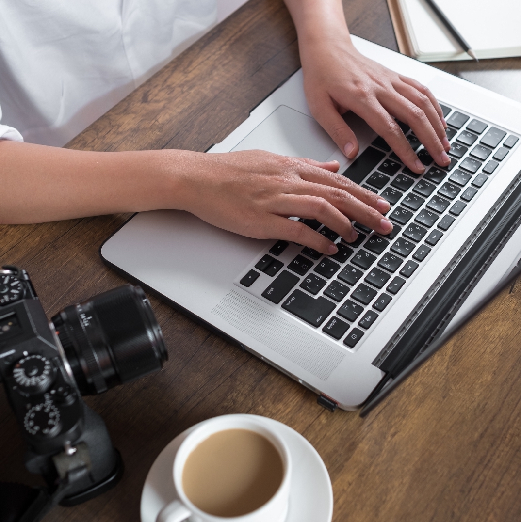 a man's hands on the laptop keyboard to preparing the hotel photography planning and strategy with camera and a cup of coffee next to the laptop and above the brown wood desk
