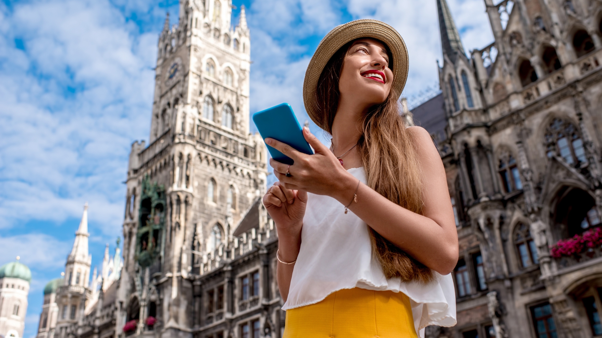 a traveller woman wear straw hat, white shirt and yellow skirt holds a blue case phone and see the landscape of historical city buildings