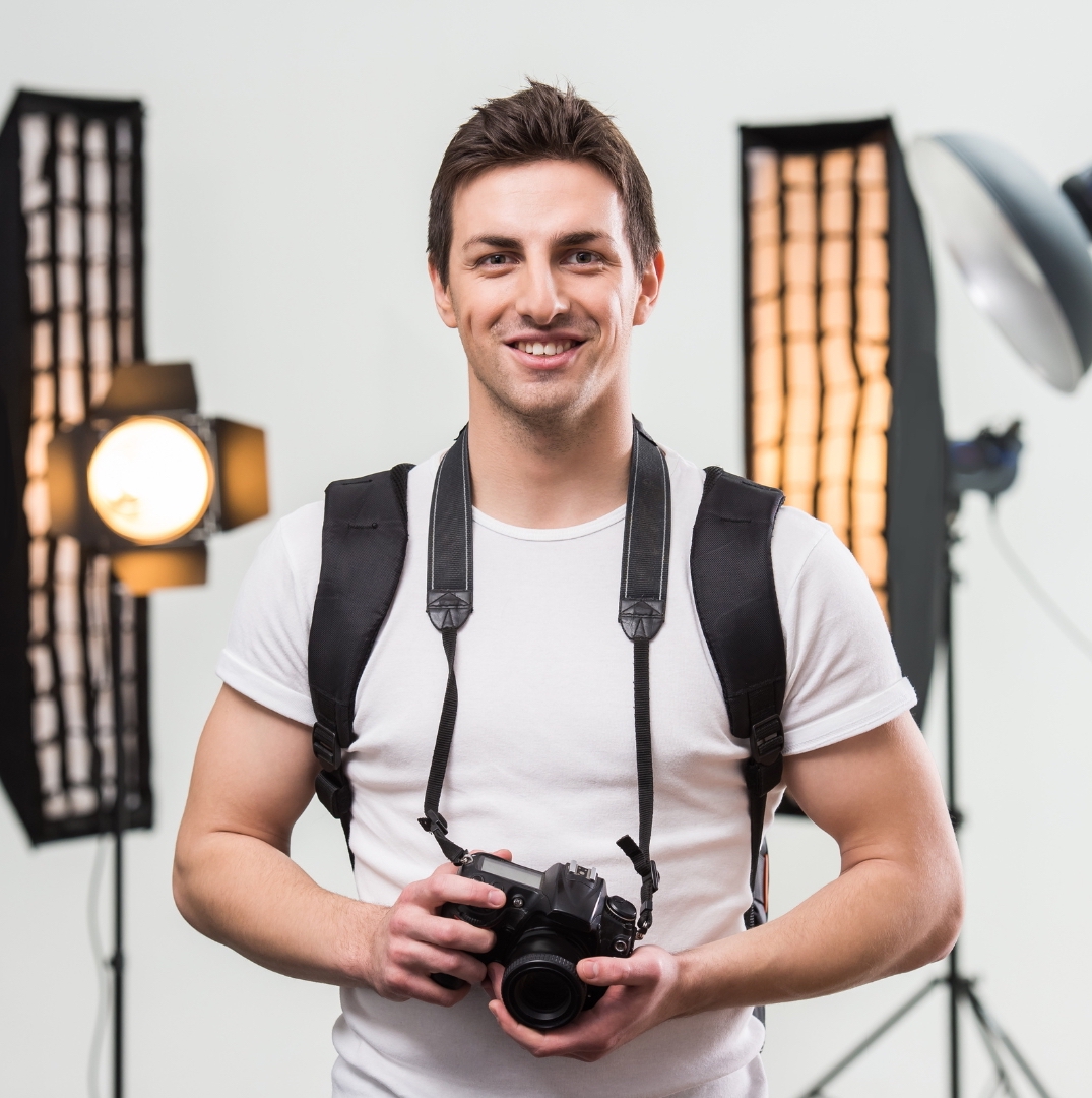 an expert architectural photographer with white shirt bring his camera and look at to the camera with two studio lightnings behind him and white wall as a background