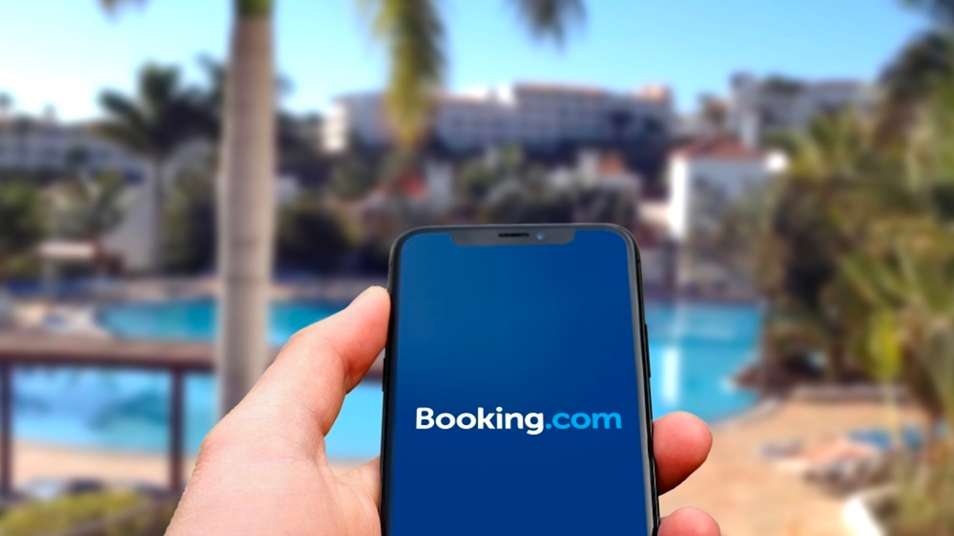 booking.com logo in white and bright blue with dark blue background on the phone that held by someone's hand with hotel landscape as a blurred background