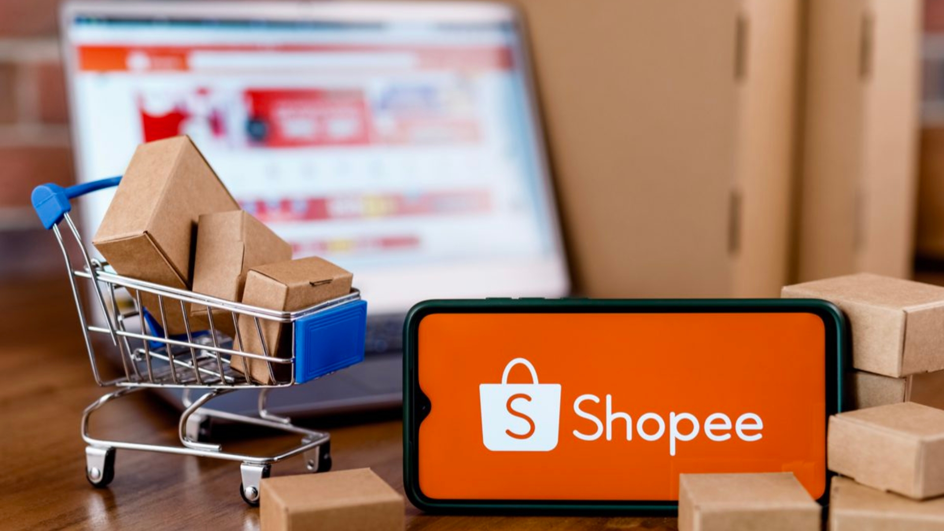 mobile phone showing shopee optimization advertising with shop trolley miniature and cardboard box inside it on the left side and laptop shows shopee website as a background