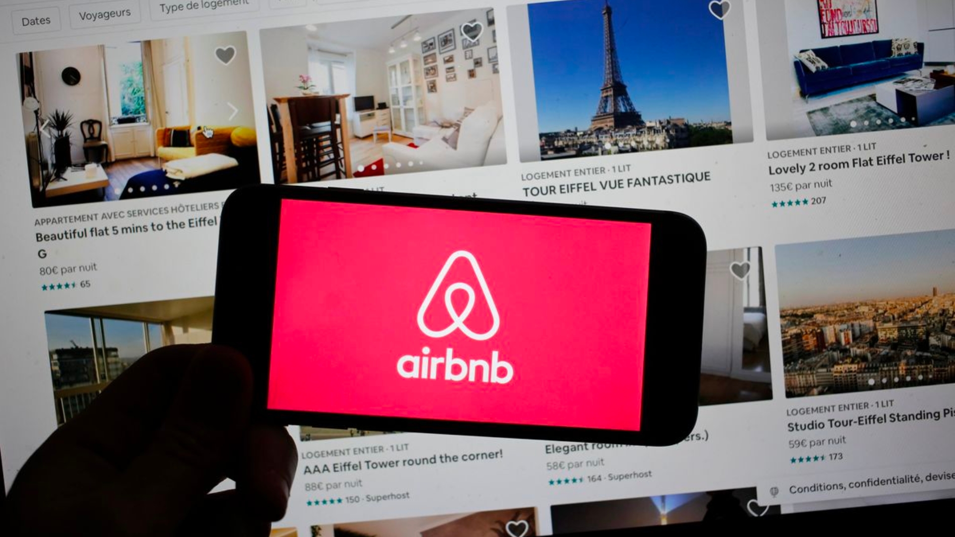 white airbnb logo with red background on the phone that held by a person's hand in silhouette and the airbnb website as a background