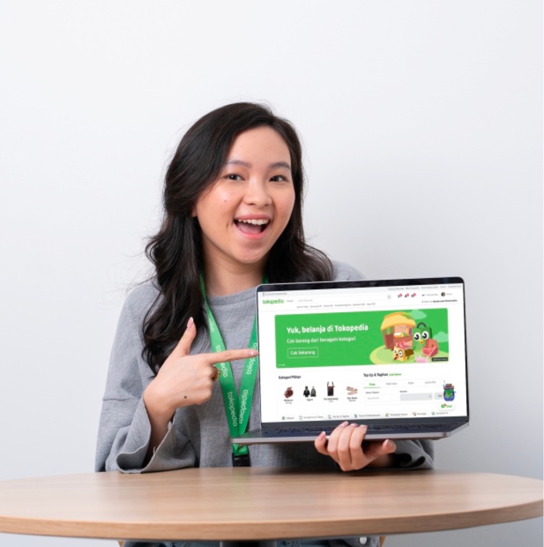 women with grey background is showing her laptop to the camera that displays tokopedia optimization